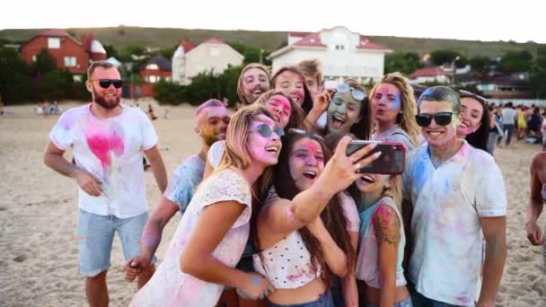 Muntre mennesker dækket i farverigt pulver tage selfie på smartphone og smil på Holi festival på stranden i slowmotion. Venner smurt ind i tørre farver tager gruppebillede på telefonen. Afslutningen på pandemien – Stock-video