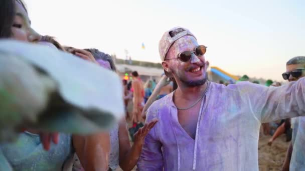 Homem sorridente leva selfie dele e amigos manchado em pó colorido no festival Holi na praia. Os amigos untados em cores secas tomam a foto de grupo pelo telefone. Fim da pandemia vívida, restrições, bloqueio — Vídeo de Stock