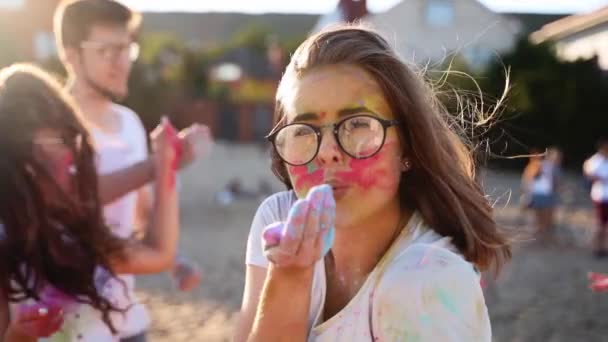 Pretty woman in glasses blows colored powder around at Holi festival on beach in slow motion. Pretty girl celebrates hindu holiday with friends. End of coronavirus quarantine, restrictions. — Stock Video