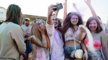 Neşeli insanlar renkli pudra atar, akıllı telefondan selfie çeker ve Holi festivalinde yavaş çekimde gülümserler. Kuru renklerde lekelenmiş arkadaşlar telefonda grup fotoğrafı çekerler. Covid salgınının sonu
