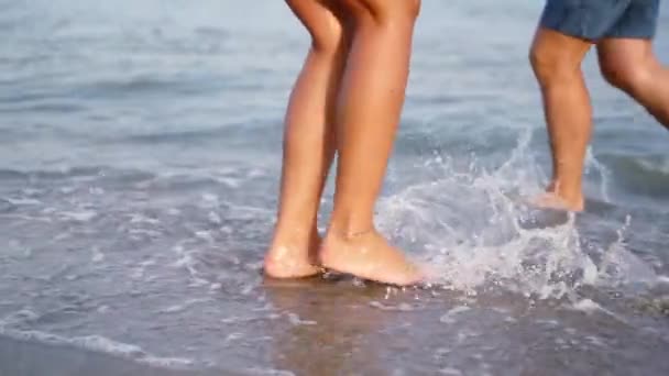 Para ucieka, tańczy wzdłuż linii surfingowej. Szczęśliwi młodzi ludzie bawią się latem na piaszczystej plaży. Smukłe opalone nogi w falach morskich — Wideo stockowe