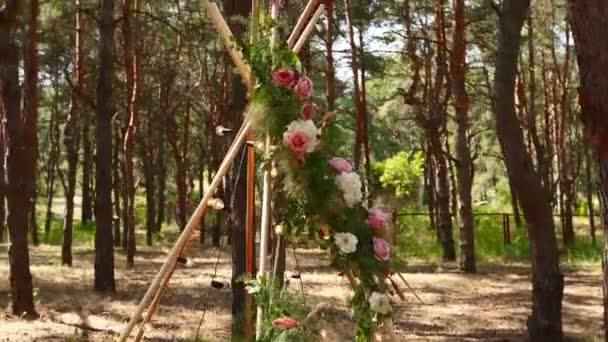 ピンクのバラで飾られた木製の棒で作られたボヘミアンティピのアーチ、カーペットの上のキャンドル、パンゲア草、松林の屋外結婚式会場の妖精の光に包まれた。写実的な構図. — ストック動画