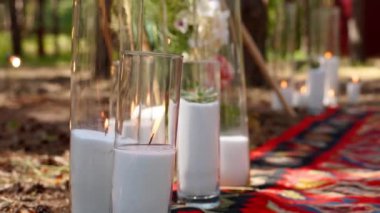 Camdan vazolarda yanan mumlar, bohem kemerinin yanındaki halının üzerinde şişeler güllerle süslenmiş ve çam ormanındaki açık hava düğün mekanında çiçeklere sarılmış çiçekler. Boho rustik dekor.