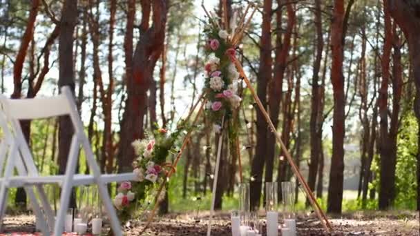Piękne cygańskie dekoracje łukowe tipi na zewnątrz miejsce ceremonii ślubnej w sosnowym lesie z szyszkami. Krzesła, kwiaciarskie kompozycje kwiatów róż, dywan, wróżkowe lampki smyczkowe. Letni ślub na wsi. — Wideo stockowe