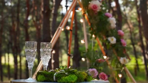 お客様のための花やキャンドルや白い椅子で飾られたbohemian tipiアーチの横にある白いテーブルの上に草や苔のワイングラス、結婚指輪の寝具. — ストック動画