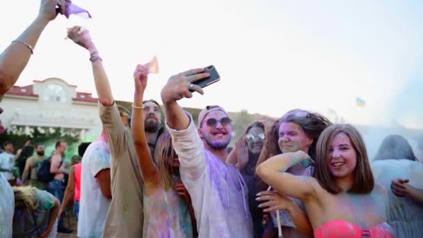 Cheerful people throw colorful powder take selfie on smartphone, jump, smile at Holi festival on beach in slow motion. Friends smeared in dry colors take group photo on phone. End of covid pandemic — Stock Video