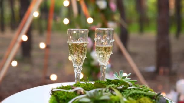 Ποτήρια κρασιού, δαχτυλίδια γάμου κλινοσκεπάσματα από γρασίδι και βρύα σε λευκό τραπέζι δίπλα στην μποέμικη αψίδα tipi διακοσμημένα σε boho στυλ με λουλούδια και κεριά και λευκές καρέκλες για τους επισκέπτες. — Αρχείο Βίντεο