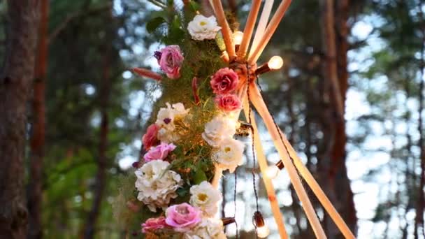 Bohemian tipi dřevěný oblouk zdobený růžovými růžemi, svíčkami na koberci, pampass trávou, zabalený v pohádkových světlech na venkovním svatebním místě v borovém lese v noci. Květinové skladby. — Stock video