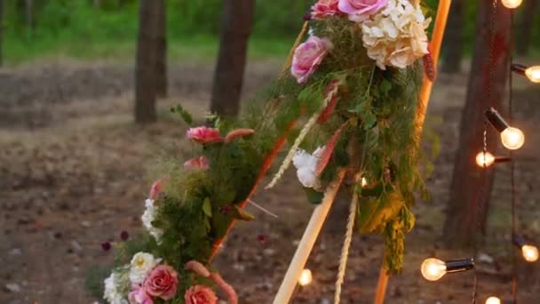 Arc en bois de tipi bohème décoré de roses roses, bougies sur tapis, herbe de pilon, enveloppé dans des lumières de fée sur le lieu de cérémonie de mariage en plein air dans la forêt de pins la nuit. Compositions floristiques. — Video
