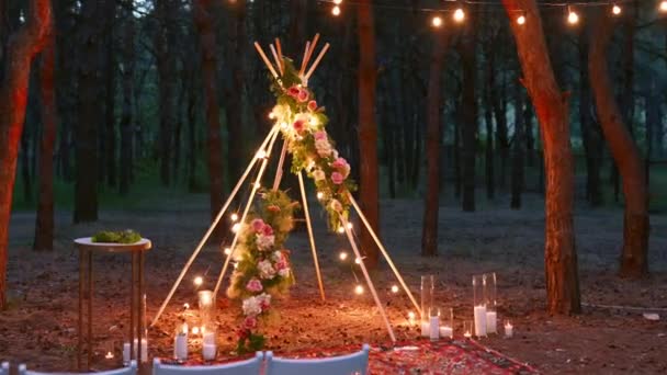 Bohemian tipi ξύλινη αψίδα διακοσμημένη με αναμμένα κεριά, τριαντάφυλλα και pampass γρασίδι, τυλιγμένη σε φώτα νεράιδα φωτισμού σε υπαίθριο χώρο τελετής γάμου στο πευκοδάσος τη νύχτα. Λαμπτήρες γιρλάντα λάμπει. — Αρχείο Βίντεο