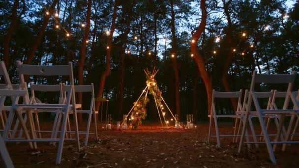 Festive lumières à cordes illumination sur boho tipi arche décor sur lieu de cérémonie de mariage en plein air dans la forêt de pins la nuit. Guirlandes d'ampoules à cordes vintage brillant au-dessus des chaises au mariage rural d'été. — Video