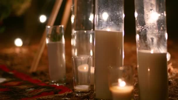 Kaarsen branden in glazen vazen, flessen op tapijt in de buurt van Boheemse tipi boog versierd met rozen en bloemen gewikkeld in feeënlichtjes op outdoor bruiloft locatie in dennenbos. Boho rustieke inrichting. — Stockvideo