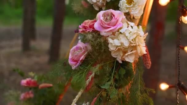 Arc en bois de tipi bohème décoré de roses roses, bougies sur tapis, herbe de pilon, enveloppé dans des lumières de fée sur le lieu de cérémonie de mariage en plein air dans la forêt de pins la nuit. Compositions floristiques. — Video