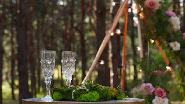 Vinglasögon, vigselringar sängkläder av gräs och mossa på vitt bord bredvid bohemisk tipi båge dekorerad i boho stil med blommor och ljus och vita stolar för gäster. — Stockvideo