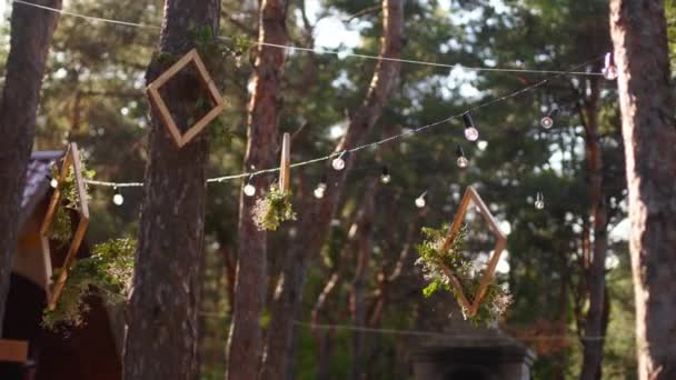 Wooden frames with floral compositions arrangement hanging on boho style wedding party venue outdoors in pine forest with string lights or fairy lights. Country or rustic eco friendly organic decor. — Stock Video