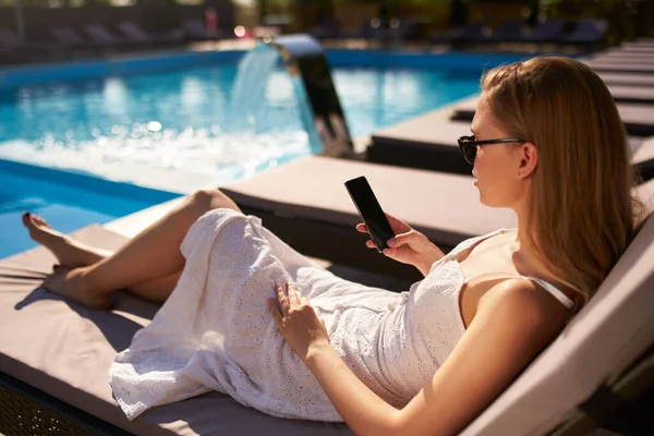 Гарненька блондинка лежить на сонці на пляжному ліжку зі смартфоном у руці біля басейну на розкішному спа-центрі. Дівчина відпочиває на стільці на тропічному курорті і використовує телефон, щоб викликати додому . — стокове фото