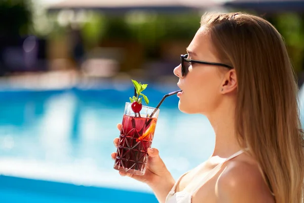 Blonde Frau trinkt erfrischenden Cocktail Sonnenbaden und sitzt in der Nähe des Swimmingpools im tropischen Spa. Frauen mit Sonnenbrille genießen einen Drink am Pool. Mädchen chillt im Urlaub in tropischem Resort. — Stockfoto