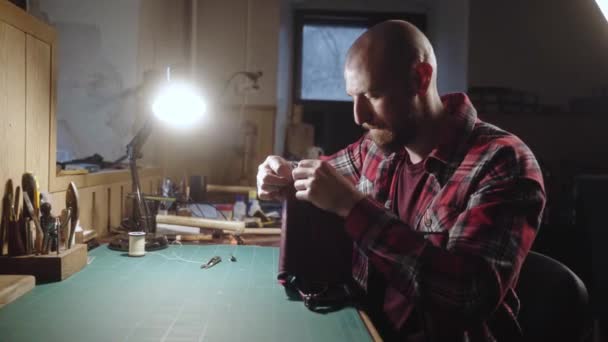 Craftsman de couro fazendo bolsa. Homem com uma agulha e linha funciona na oficina de couro. Alfaiate em camisa xadrez fazendo uma bolsa de costura acessórios no atelier. — Vídeo de Stock