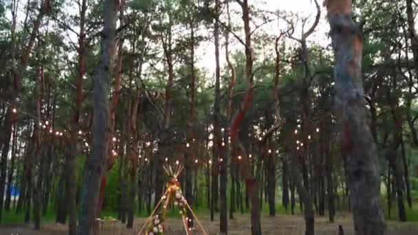 Ünnepi húros fények világítás boho tipi boltív dekoráció szabadtéri esküvői helyszín fenyves erdőben éjszaka. Vintage húr lámpák izzók koszorúk ragyog a székek felett a nyári vidéki esküvő. — Stock videók
