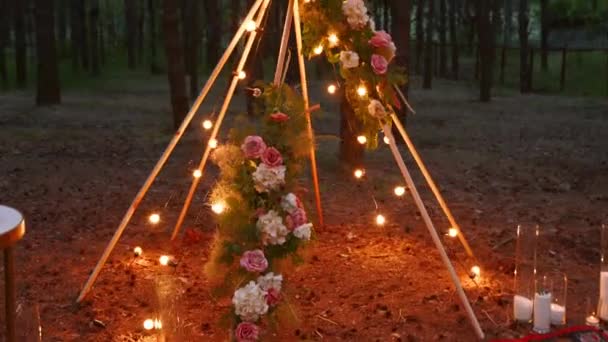 Arco de madera de tipi bohemio decorado con velas encendidas, rosas y hierba de pasto, envuelto en luces de hadas iluminación en el lugar de la ceremonia de boda al aire libre en el bosque de pinos por la noche. Bulbos guirnalda brilla. — Vídeos de Stock