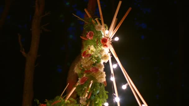Arc en bois de tipi bohème décoré avec des bougies allumées, des roses et de l'herbe pampass, enveloppé dans des lumières de fées illumination sur le lieu de cérémonie de mariage en plein air dans la forêt de pins la nuit. Bulbes guirlande brille. — Video