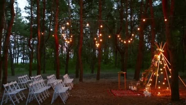 Festlig sträng ljus belysning på boho tipi båge inredning på utomhus bröllop plats i tallskog på natten. Vintage sträng lampor glödlampa girlanger lyser ovan stolar på sommaren lantligt bröllop. — Stockvideo