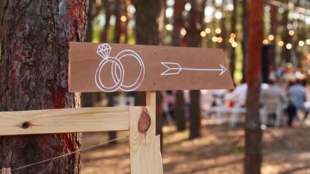 Panah kayu tanda dengan cincin pernikahan di tempat upacara. Pos panji pesta pernikahan. Banner informasi Arah untuk tamu di hutan pinus luar ruangan. Dekorasi gaya pedesaan atau pedesaan. — Stok Video