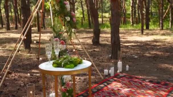 Bohemian tipi arch made of wooden rods decorated with pink roses, candles on carpet, pampass grass, wrapped in fairy lights on outdoor wedding ceremony place in pine forest. Květinové skladby. — Stock video
