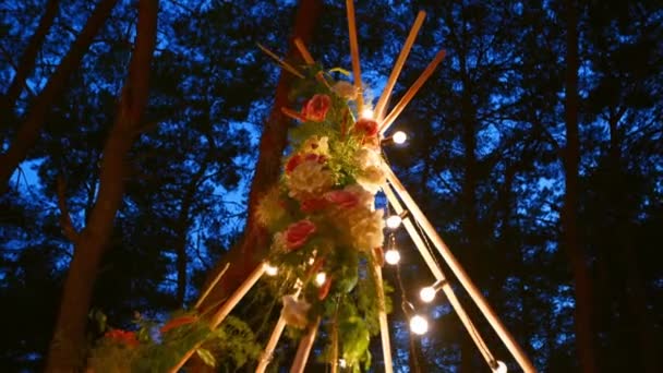Bohemian tipi dřevěný oblouk zdobený hořícími svíčkami, růžemi a pampass trávou, zabalený v pohádkovém osvětlení na venkovním svatebním obřadu v borovém lese v noci. Žárovky svítí. — Stock video