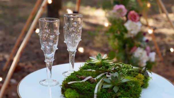 Copas de vino, anillos de boda ropa de cama de hierba y musgo en mesa blanca junto al arco de tipi bohemio decorado en estilo boho con flores y velas y sillas blancas para invitados. — Vídeos de Stock