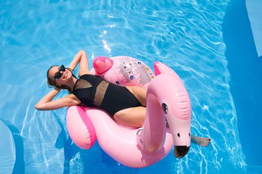 Yüzme havuzunda şişme pembe flamingo şilteli bikinili seksi kadın dinleniyor. Mayo giymiş çekici bir kız güneşin altında yüzer vaziyette yatıyor. Güzel bayan tropik tatilde dinleniyor..