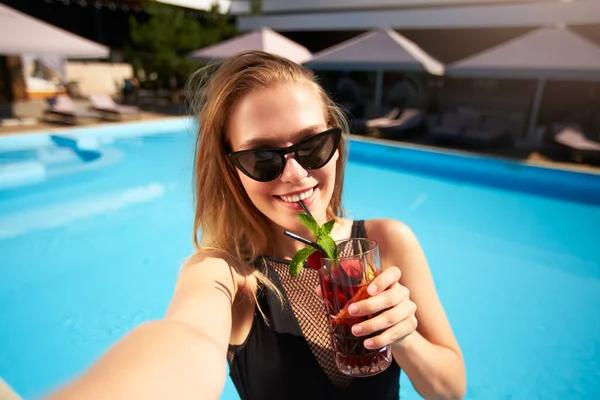 Travel blogger woman in bikini taking selfie photo with cocktail near swimming pool. Pov of lifestyle vlogger selfie from vacation on luxury resort. Female in swimwear drinks beverage on tropical spa. — ストック写真