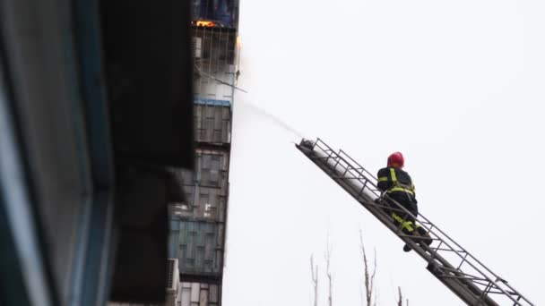 Un pompier sur un escalier aérien éteint un grand incendie dans un immeuble résidentiel de plusieurs étages. Tuyau de pompier vers le bas des flammes sur une échelle à plateau tournant élevé. — Video