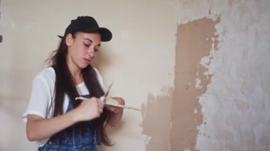 Profesyonel alçı dişisi duvarı alçılıyor, alçıyı tulum ve beyzbol şapkasıyla bir spatulaya sürüyor ve yayıyor. Duvarları sıva kaplı genç bir kadın, inşaat alanında dekorasyon işi yapıyor. İşlemler.