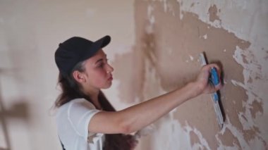 Profesyonel alçı dişisi duvarı alçılıyor, alçıyı tulum ve beyzbol şapkasıyla bir spatulaya sürüyor ve yayıyor. Duvarları sıva kaplı genç bir kadın, inşaat alanında dekorasyon işi yapıyor. İşlemler.