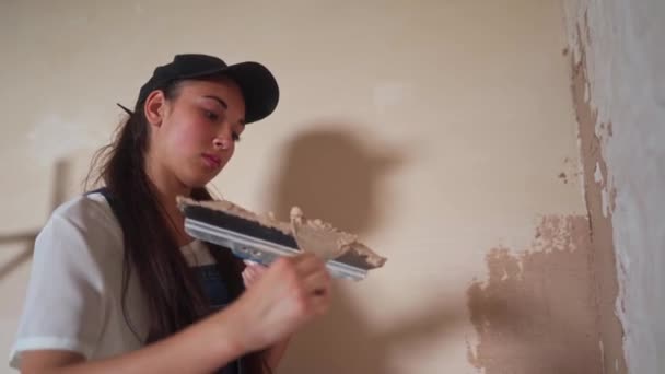 Profesional yesero hembra salpica la pared, se aplica y se extiende yeso en una espátula en overoles y gorra de bola. Joven mujer paredes de yeso, haciendo trabajos de decoración en el sitio de construcción. Ocupaciones. — Vídeo de stock