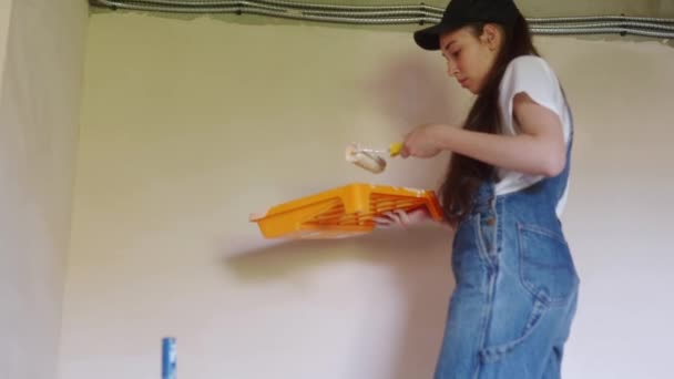 Professionell målare hona målar väggen med rulle och bricka på byggarbetsplatsen. Ung kvinna i overaller och mössa gör heminredning och renovering. Arbetstagare gör gör DIY reparationsarbete i rummet. — Stockvideo
