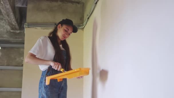 La pintora profesional pinta la pared con rodillo y bandeja en el sitio de construcción. Mujer joven en overoles y gorra haciendo decoración y renovación del hogar. Trabajador hace trabajos de reparación de bricolaje en la habitación. — Vídeo de stock