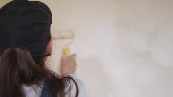 Profesyonel ressam kadın, inşaat alanında duvarı silindir ve tepsiyle boyuyor. Tulumlu ve şapkalı genç bir kadın ev dekorasyonu ve tadilat yapıyor. İşçi odada DIY onarımı yapıyor. — Stok video