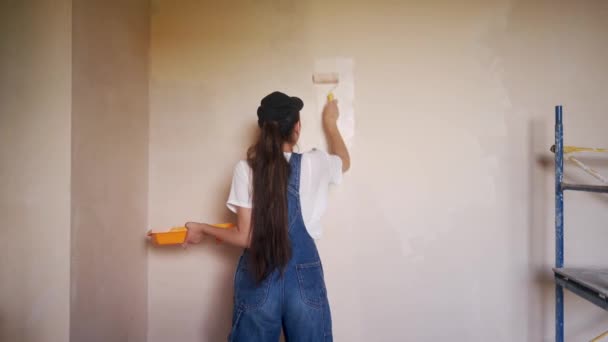 Profissional pintor feminino pinta a parede com rolo e bandeja no canteiro de obras. Jovem mulher de macacão e boné fazendo decoração e renovetion casa. Trabalhador faz trabalhos de reparação DIY na sala. — Vídeo de Stock