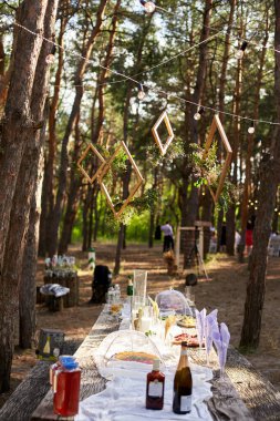Düğün partisi dışarıda ormanda ziyafet veriyor. Mum, beyaz kumaş, çiçek, tabak, sofra takımı, yemek takımı, yemek takımı, kadeh ve içeceklerle süslenmiş yemek masası. Tatil yemekleri..