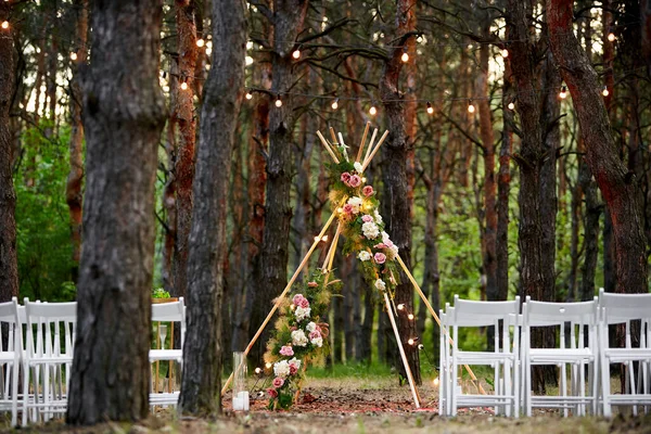 Piękne cygańskie dekoracje łukowe tipi na zewnątrz miejsce ceremonii ślubnej w sosnowym lesie z szyszkami. Krzesła, kwiaciarskie kompozycje kwiatów róż, dywan, wróżkowe lampki smyczkowe. Letni ślub na wsi. — Zdjęcie stockowe