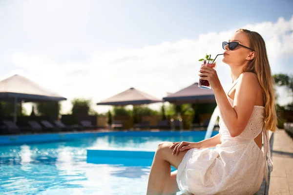 Блондинка п'є освіжаючий коктейль засмагає і сидить біля басейну в тропічному спа-центрі. Жінка в сонцезахисних окулярах насолоджується випивкою на березі басейну. Дівчина охолоджує в тропічному курорті у відпустці . — стокове фото