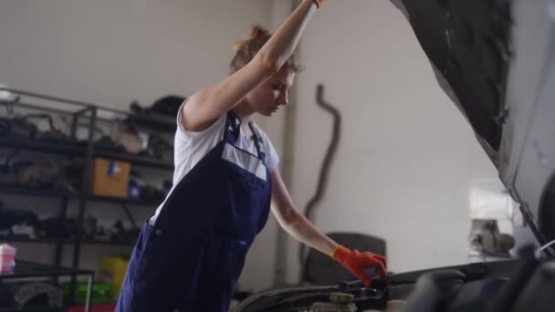 Kadın tamirci teftiş için araba kaputunu açar. Tulumlu ve koruyucu eldivenli kadın araba tamir servisindeki motor ve parçaları kontrol ediyor. Modern atölyede araç tamiri hatası. — Stok video