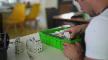 Elektrikçi, elektrik paneli sigorta kutusunu atölyede monte ediyor. İşçi masadaki dağıtım panosuna enerji sayacı, şalterler ve devre kesiciler kurar.