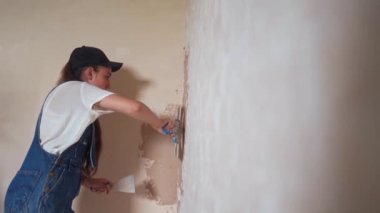 Profesyonel alçı dişisi duvarı alçılıyor, alçıyı tulum ve beyzbol şapkasıyla bir spatulaya sürüyor ve yayıyor. Duvarları sıva kaplı genç bir kadın, inşaat alanında dekorasyon işi yapıyor. İşlemler.