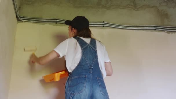 Professionelle Malerin malt die Wand mit Walze und Tablett auf der Baustelle. Junge Frau in Overalls und Mütze beim Dekorieren und Renovieren. Arbeiter macht Reparaturarbeiten im Zimmer. — Stockvideo