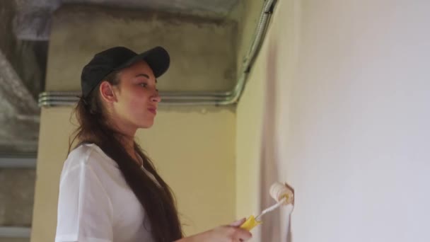 Profi festő nő festi a falat görgővel és tálcával az építési területen. Fiatal nő overálban és sapkában, lakberendezéssel és felújítással. Munkás teszi DIY javítási munka a szobában.