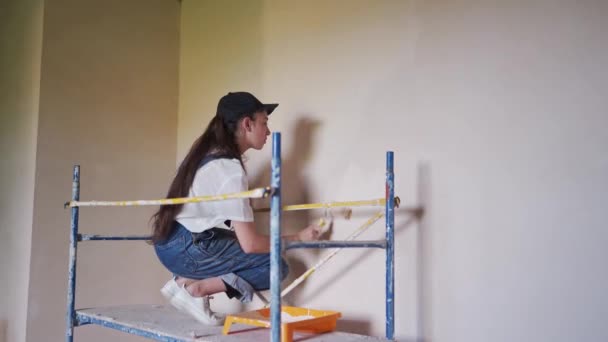 Female painter paints wall with roller on scaffolding at construction site. Young woman in overalls doing home decoration, renovation on staging. Professional worker makes DIY repair work in room. — Vídeo de Stock