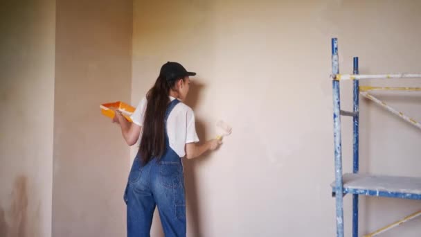 Profissional pintor feminino pinta a parede com rolo e bandeja no canteiro de obras. Jovem mulher de macacão e boné fazendo decoração e renovetion casa. Trabalhador faz trabalhos de reparação DIY na sala. — Vídeo de Stock